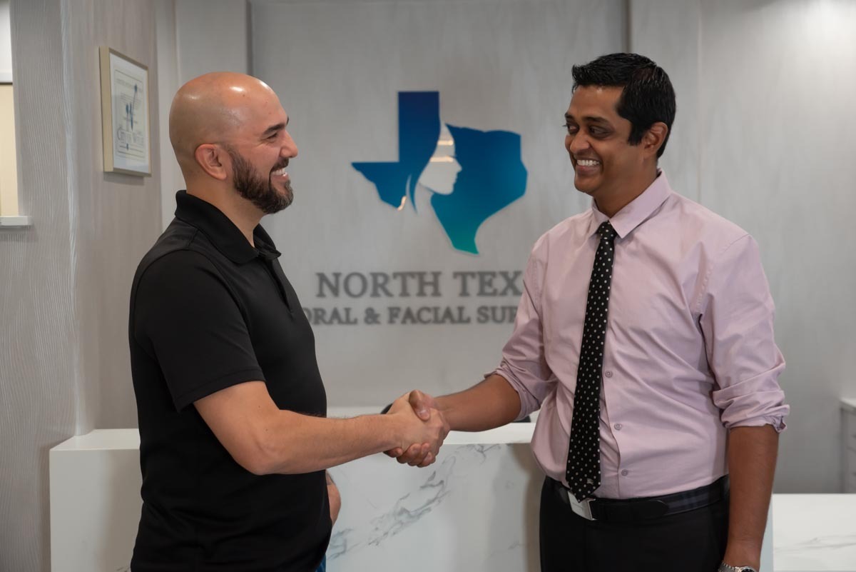Dr. Ajay Ganti shakes hands with a patient in the reception area of North Texas Oral & Facial Surgery, both smiling during their greeting.